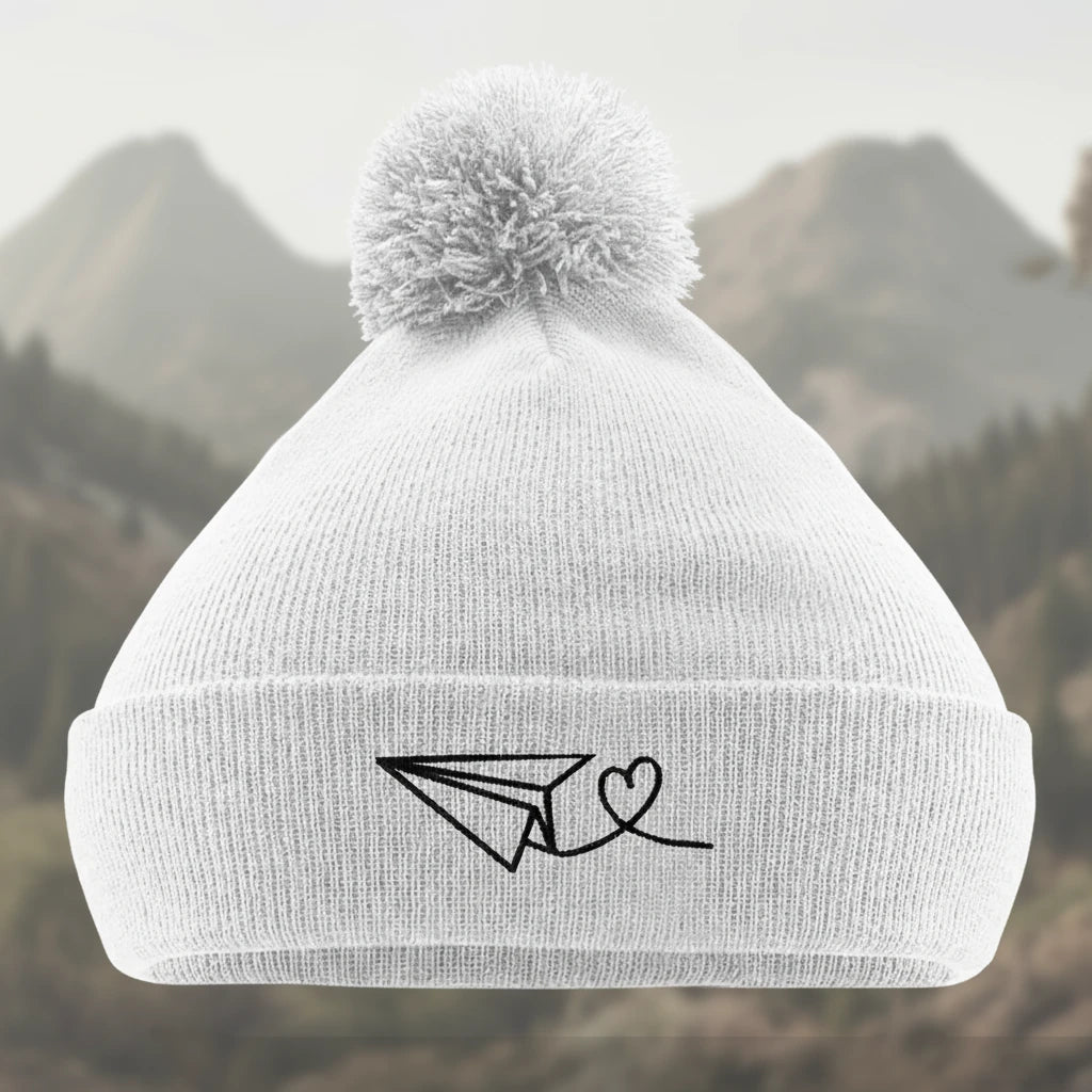 Weiße Bommelmütze mit gesticktem Papierflieger und Herz-Motiv. Symbol für Reiselust und Liebe zum Abenteuer.